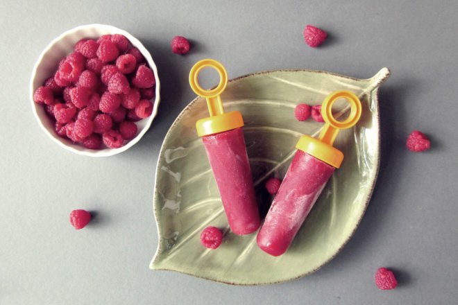two popsicles on a leaf-shaped plate near a bowl of raspberries