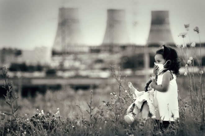 A young girl stands in a field, holding a doll and wearing a mask over her nose and mouth. In the background, there is a factory.