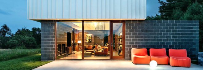 Image of the exterior of the cabin at night. A patio has a few orange lounge chairs. Large windows show the inside of the cabin, where the family is sitting on an orange sectional couch.