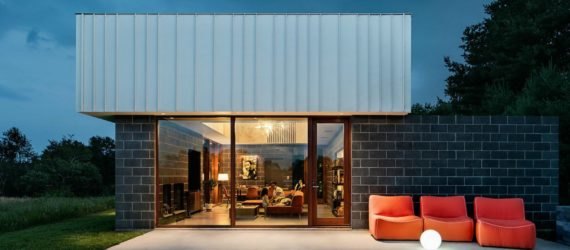 Image of the exterior of the cabin at night. A patio has a few orange lounge chairs. Large windows show the inside of the cabin, where the family is sitting on an orange sectional couch.
