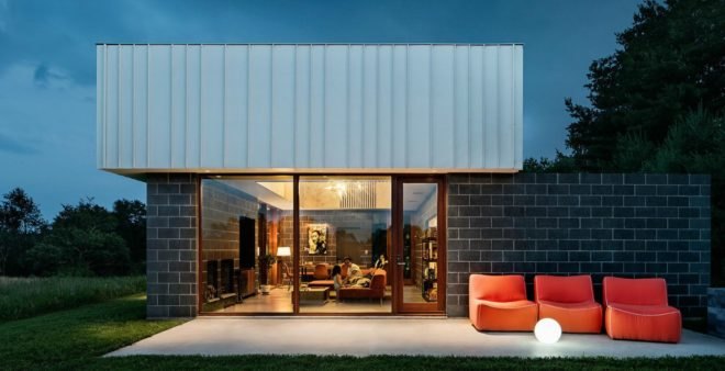 Image of the exterior of the cabin at night. A patio has a few orange lounge chairs. Large windows show the inside of the cabin, where the family is sitting on an orange sectional couch.