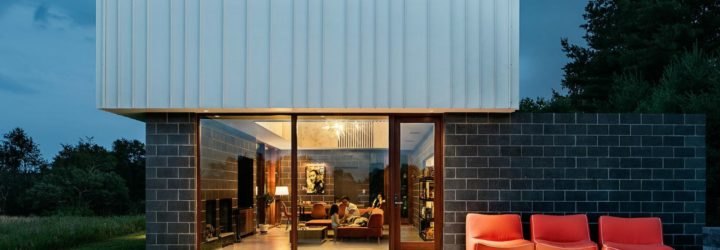 Image of the exterior of the cabin at night. A patio has a few orange lounge chairs. Large windows show the inside of the cabin, where the family is sitting on an orange sectional couch.