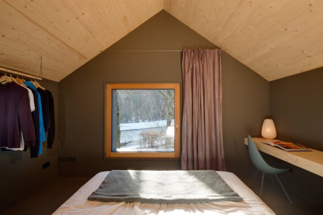 image of a bed, a square window, a wooden desk, and clothes hanging on a rack suspended from the ceiling