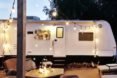 A fire pit is blazing in front of a white camper, which has been transformed into a tiny home complete with decorative string lights.