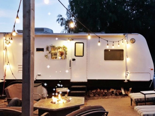 A fire pit is blazing in front of a white camper, which has been transformed into a tiny home complete with decorative string lights.