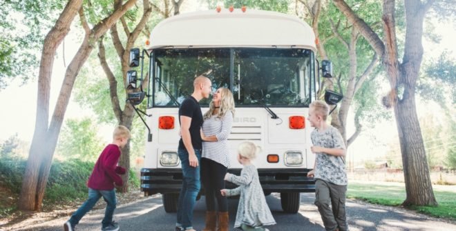 Brandon and Ashley Trebitowski look at each other while their three children run around them. The family is in front of a white bus that they converted into their tiny home.