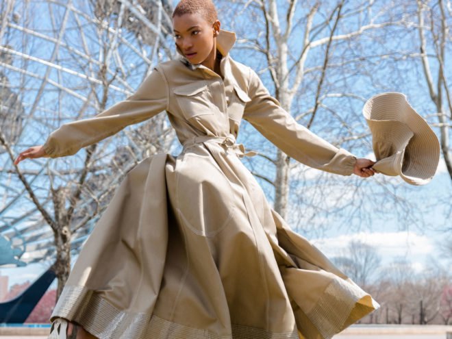 Model twirls in a tan trench coat and holds a tan hat, both items made from recycled water bottles.