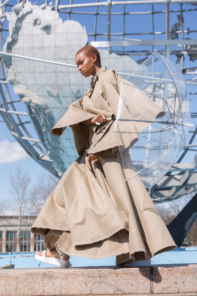 Model in a tan trench coat holds a clear umbrella.