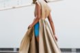 Model walks wearing a tan trench coat with tall leg slits and sports bright blue shorts underneath. The trench connects to a hat.