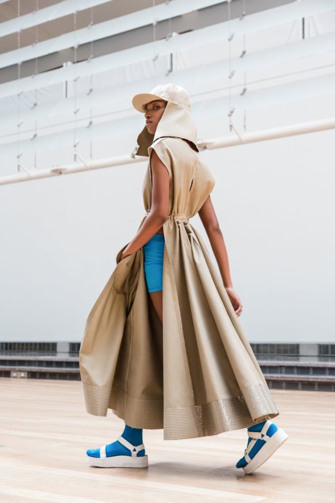 Model walks wearing a tan trench coat with tall leg slits and sports bright blue shorts underneath. The trench connects to a hat.