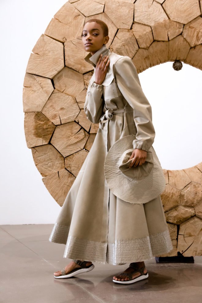 Model wears trench coat and holds a hat.