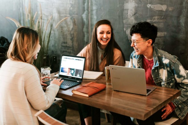 Three people are sitting at a table, hunkered over their laptops and laughing.