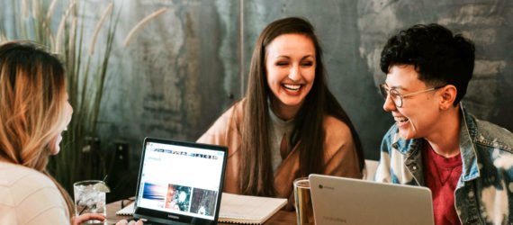 Three people are sitting at a table, hunkered over their laptops and laughing.
