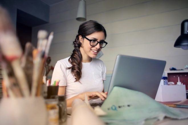 A woman with glasses sits at a desk and types on a laptop.