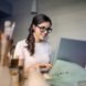 A woman with glasses sits at a desk and types on a laptop.