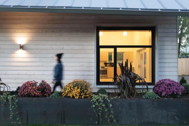 An out-of-focus woman walking past a white clapboard facade and lit-up window