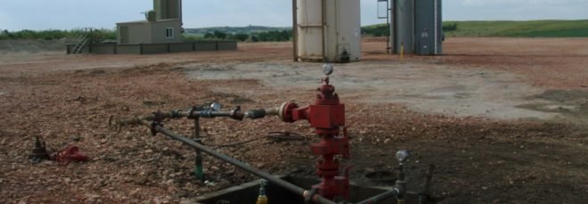 A red fracking well head with abandoned buildings in the background