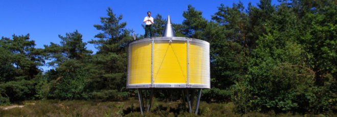 man standing on top of round yellow pavilion on metal supports in forest landscape