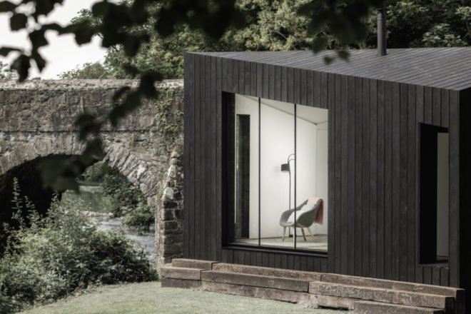 black cabin with large glass window by a stone bridge