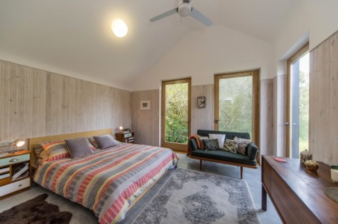 Bedroom with colorful striped bedding, couch with several throw pillows, double height ceilings and large windows with views of woodlands