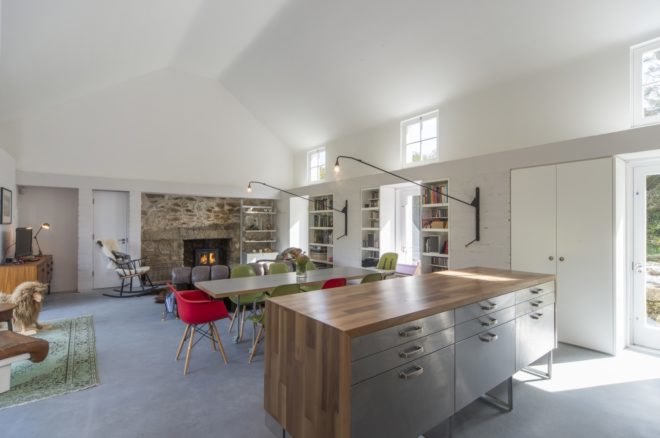 Open-plan living area with kitchen island in foreground, long dining table with green and red chairs in midground and gray couch and rocking chair facing a fireplace in background