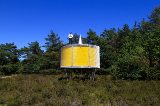 man standing on top of round yellow pavilion on metal supports in forest landscape