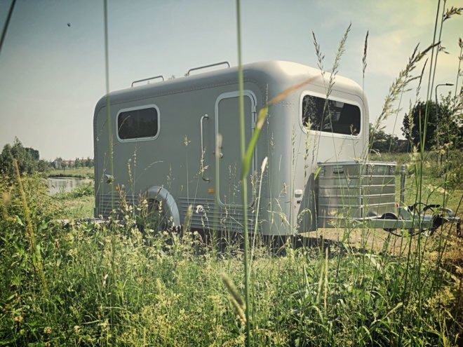 Field in foreground and aluminum trailer parked near a lake