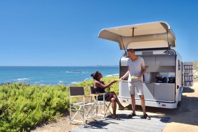 Two people sharing food near trailer's outdoor kitchen with beach in the background