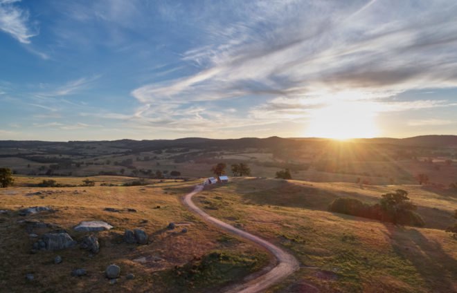 aerial view of tiny brick cabin and white shed on vast landscape of rolling hills at sunset