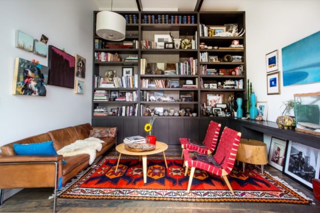 floor-to-ceiling dark wood bookshelf filled with books and small knickknacks and a tan leather couch and red chairs on a colorful patterned rug