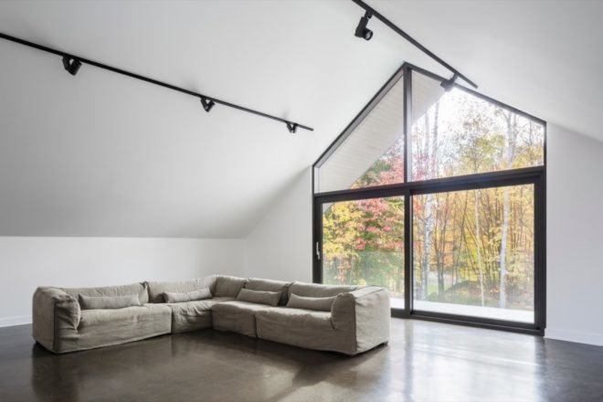 large white room with pitched ceiling, tan sectional sofa, and glazed wall facing the forest