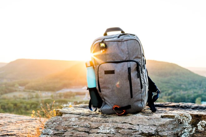 gray backpack with blue water bottle in side pocket sitting on a rock with sunset in background