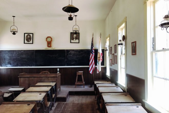 Empty classroom with wooden desks, wooden teacher desk at front of room and black chalkboard