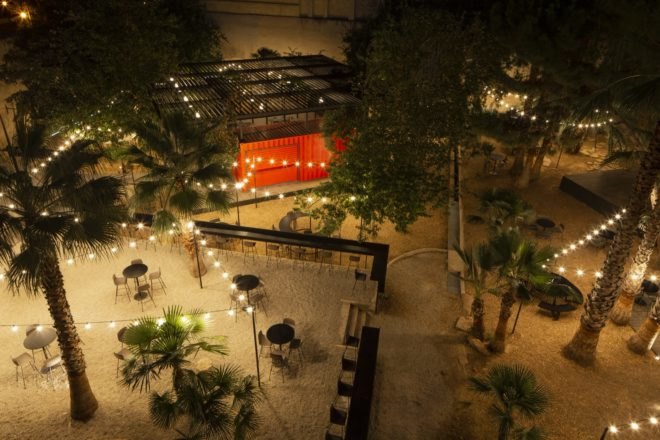 aerial view of outdoor dining area with palm trees, black tables and chairs, twinkle lights and a red shipping container