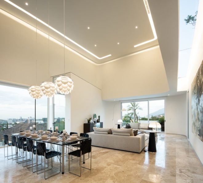 white room with double-height ceiling and glass walls, beige couches and a long glass dining table with black chairs