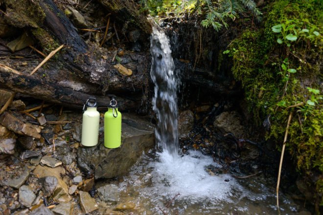two reusable water bottles on a rock in front of a waterfall