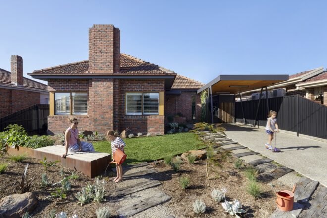 woman sits on garden wood box while children lay and water plants near drive way