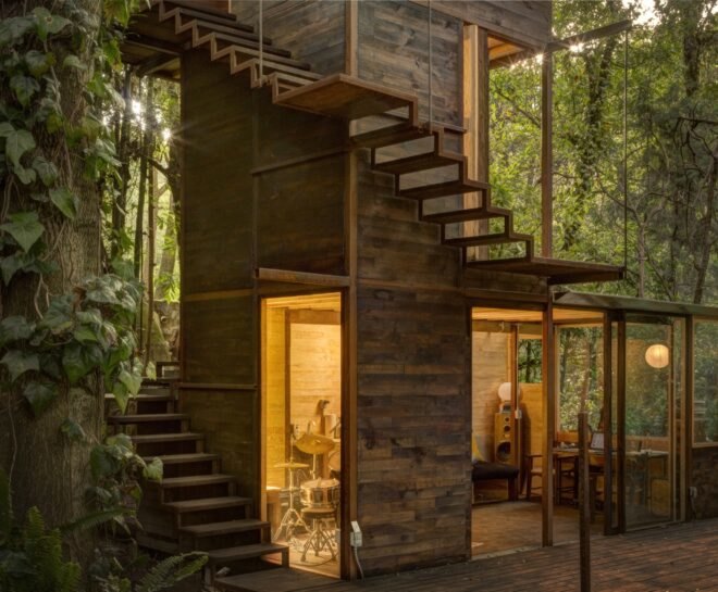 wood treehouse with wood spiraled stairs surrounded by forest with interior lights illuminating the home