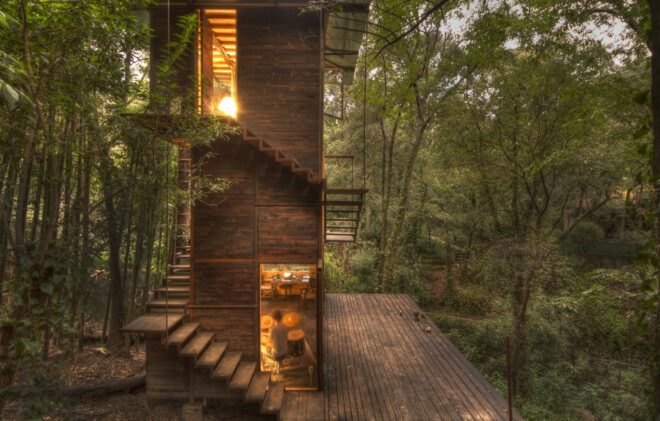 wood treehouse with wood spiraled stairs surrounded by forest with interior lights illuminating the home