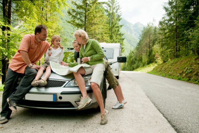 a family sits on a car's hood and look at map while on the road neat a forest
