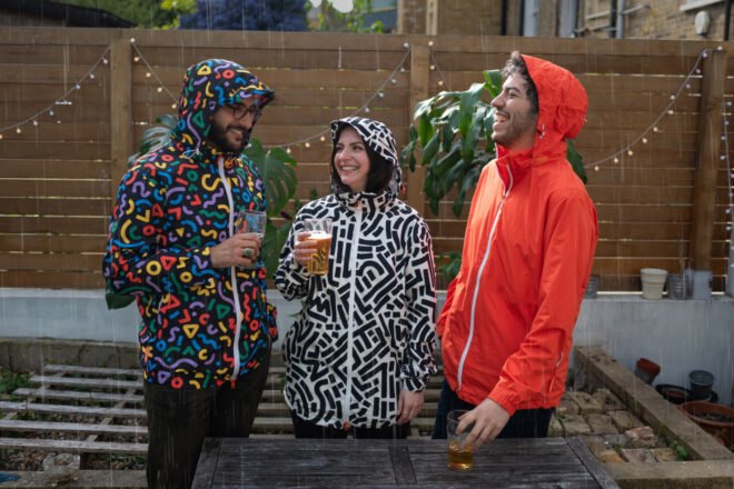 three friends in bright and patterned rain jackets drinking beer in the rain