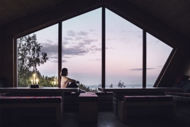 person sitting on bench near large window overlooking fjords at sunset