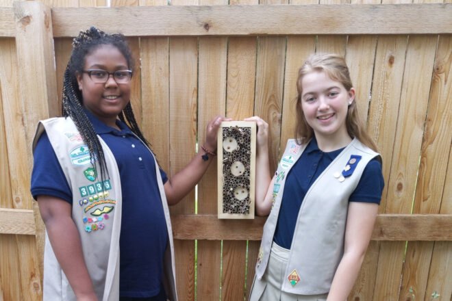 two Girl Scouts members smiling next to a handmade bee hotel on a fence