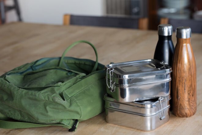 green backpack, two tin lunch boxes and a two reusable water bottles on a wood table