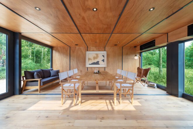 interior of building with wood floors, walls and ceilings. a long table with chairs sits in the middle of the room, with windows on both the left and right of the room. to the left is a blue sofa.