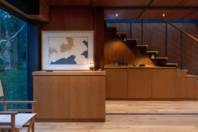 to the left is a floor-to-ceiling window. next to the window is a wooden cabinet with a framed artwork hanging above it. to the right is another cabinet and a set of stairs.