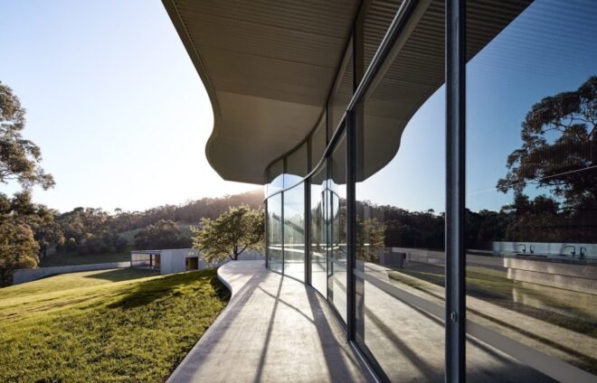 a long view of sunlight glinting off the horizontal building's floor-to-ceiling windows. in the background is another building made of cement, with grass and trees surrounding it
