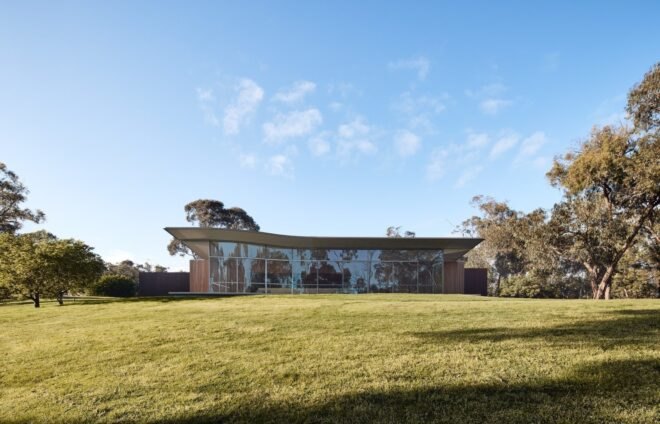 a long horizontal building with a wall of floor-to-ceiling windows, a green lawn in front of the building and trees on both sides of the building