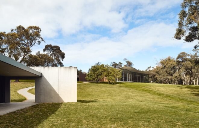 a long view of the horizontal building with a gray wall to the left in the foreground and trees next to the building in the background. the gray building in the foreground has an open walk-through area.