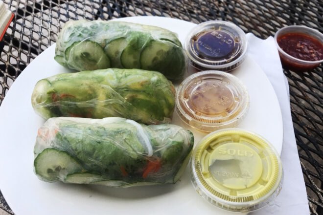 A plate with three green spring rolls and three small cups of sauces to the right, with a fourth cup of sauce on the table